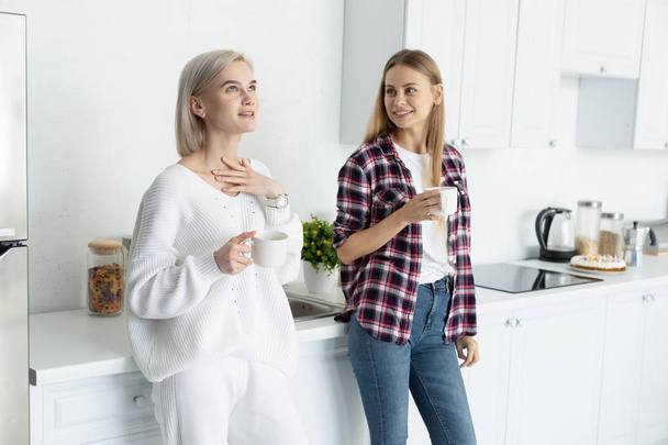 Two young beautiful females in casual clothes spending time together in the kitchen - Photo, Image