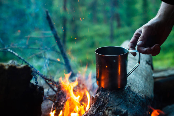 Adam şenlik ateşinde metal kupada çay ısıtıyor. Doğada sıcak içecek. Açık havada çay içmek. İnsan elinde çelik kupa. Gün batımında kamp. Alacakaranlıkta romantik sıcak atmosfer açık . Aktif dinlenme. - Fotoğraf, Görsel