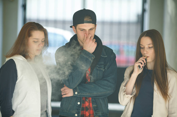 Vape teenagers. Two young cute girls in casual clothes smoke electronic cigarettes near nonsmoking guy outdoors in the street in summer day. Bad habit that is harmful to health. Vaping activity. - Fotoğraf, Görsel
