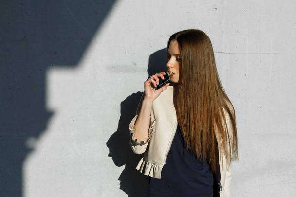 Vape teenager. Young cute girl in  casual clothes smokes an electronic cigarette near the wall outdoors in summer day. Bad habit that is harmful to health. Vaping activity. Close up. - Foto, Bild