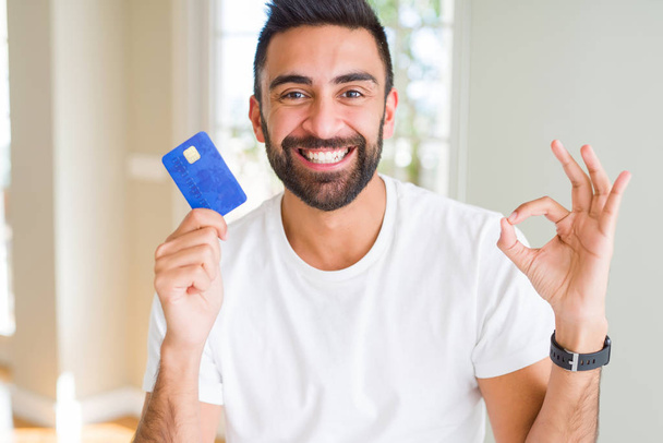 Bonito hispânico homem segurando cartão de crédito fazendo ok sinal com os dedos, excelente símbolo
 - Foto, Imagem