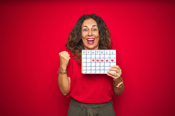 Старша жінка середнього віку тримає календар менструацій над червоним ізольованим фоном, кричить гордо і святкує перемогу і успіх дуже схвильовані, веселі емоції
 - Фото, зображення