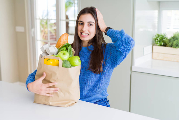 Beautiful young woman holding paper bag full of healthy groceries stressed with hand on head, shocked with shame and surprise face, angry and frustrated. Fear and upset for mistake. - Photo, Image