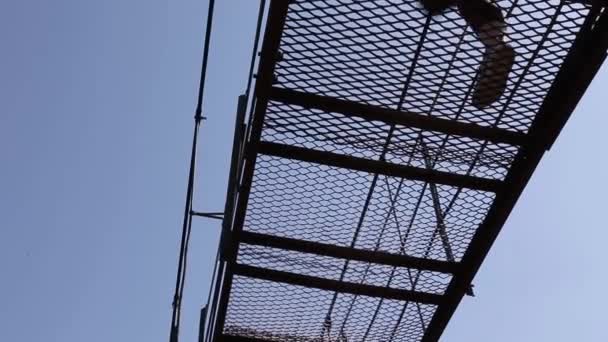 People walk on a wobbly metal lattice suspension bridge. View from the bottom through the perforated floor against the blue sky. - Footage, Video