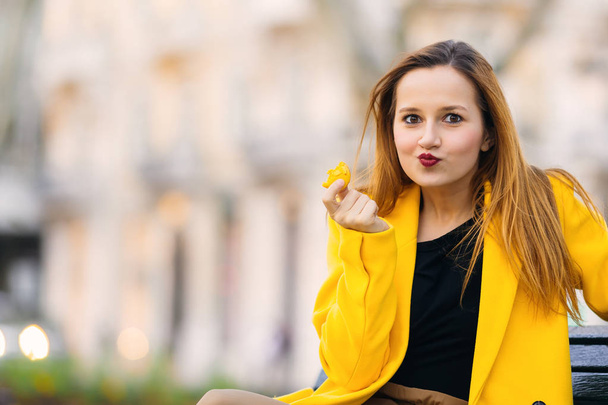 Mädchen in gelbem Mantel sitzt auf der Bank und genießt eine Delikatesse - Foto, Bild