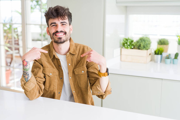 Jongeman draagt casual jas zittend op witte tafel zelfverzekerd kijkend met een glimlach op het gezicht, wijzend met de vingers trots en gelukkig. - Foto, afbeelding