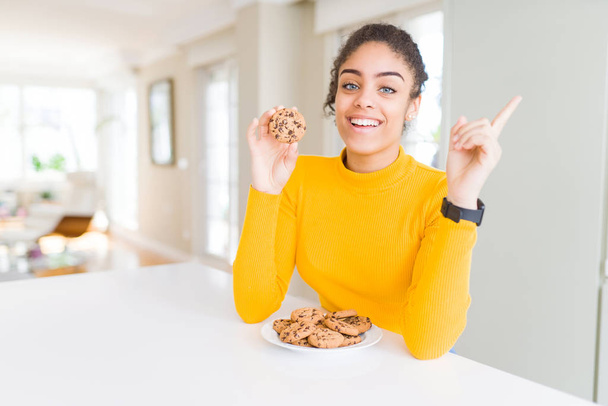 Молода афроамериканська дівчинка, яка їсть шоколадні тістечка, як солодка закуска, дуже щаслива, показуючи пальцем на бік. - Фото, зображення