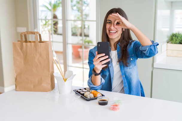 Bella giovane donna che ordina la consegna del cibo da app utilizzando smartphone con il viso felice sorridente facendo ok segno con mano sugli occhi guardando attraverso le dita
 - Foto, immagini