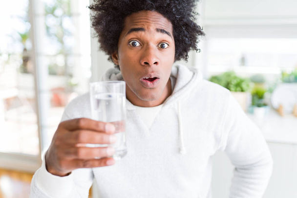 Hombre afroamericano bebiendo un vaso de agua en casa asustado en shock con una cara sorpresa, asustado y excitado con la expresión del miedo
 - Foto, imagen