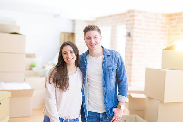 Όμορφο νεαρό ζευγάρι που κινείται σε ένα νέο σπίτι, στέκεται σε νέο διαμέρισμα γύρω από χαρτοκιβώτια, αγκαλιάζοντας χαμογελαστά ευτυχισμένοι και ερωτευμένοι - Φωτογραφία, εικόνα