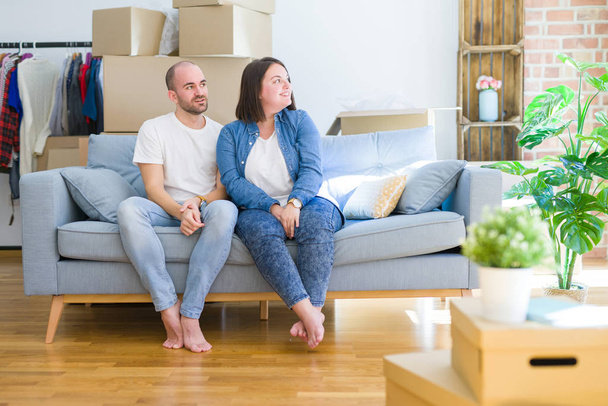 Young couple sitting on the sofa arround cardboard boxes moving to a new house looking away to side with smile on face, natural expression. Laughing confident. - Foto, immagini