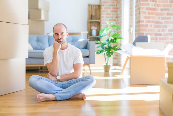 Young bald man sitting on the floor around cardboard boxes moving to a new home with hand on chin thinking about question, pensive expression. Smiling with thoughtful face. Doubt concept. - Photo, Image