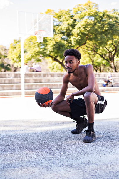 Joven atlético negro posando en la cancha con pelota, jugador de baloncesto, ejercicios matutinos, estilo de vida saludable activo, día caluroso de verano, streetball
. - Foto, imagen
