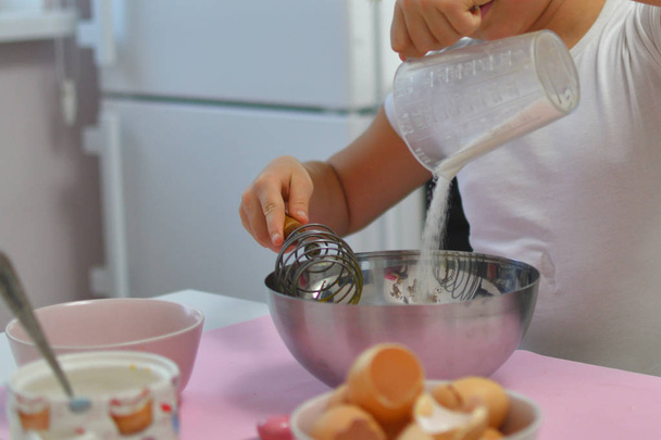 the girl pours sugar from a measuring glass in a metal bowl. Little girl preparing cookies in kitchen at home. Cooking homemade food. - 写真・画像