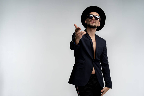 Happy attractive young bearded man in stylish black suit and hat in black sunglasses, posing with outstretched hand isolated over white background - Photo, Image
