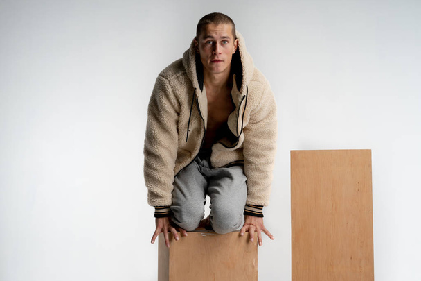 Young attractive man with short haircut, wearing sportswear, kneeling on wooden cube, looking at the camera with funny face isolated over white background - Valokuva, kuva