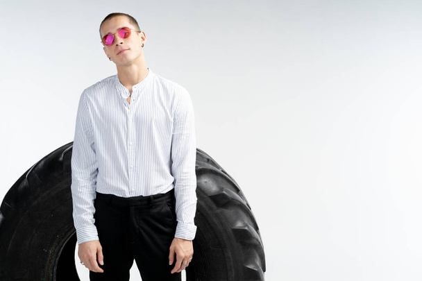 Handsome young man in white shirt in red sunglasses standing in front of the big tire, looking at the camera, isolated over white background, copyspace for your text - Foto, Imagem
