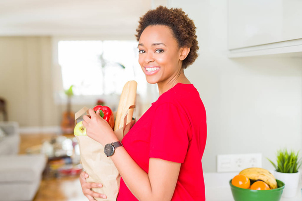 Young beautiful african american woman holding paper bag full of fresh healthy groceries at the kitchen - Foto, afbeelding
