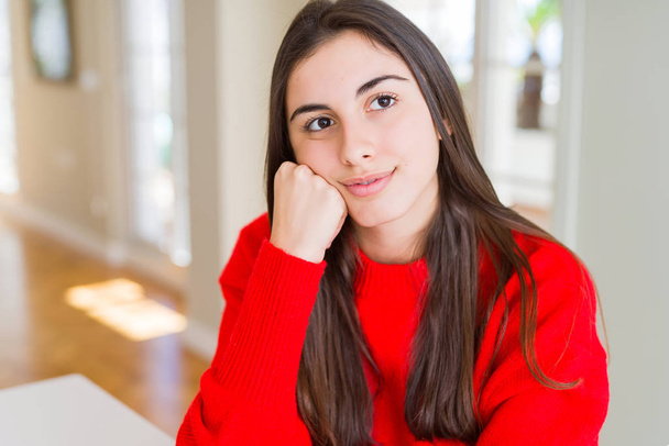 Beautiful young woman wearing casual red sweater with hand on chin thinking about question, pensive expression. Smiling with thoughtful face. Doubt concept. - Fotó, kép