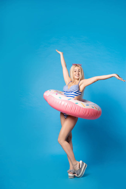 Full-length photo of happy blonde with raised hands in bathing suit with inflatable donut on empty blue background - Foto, imagen