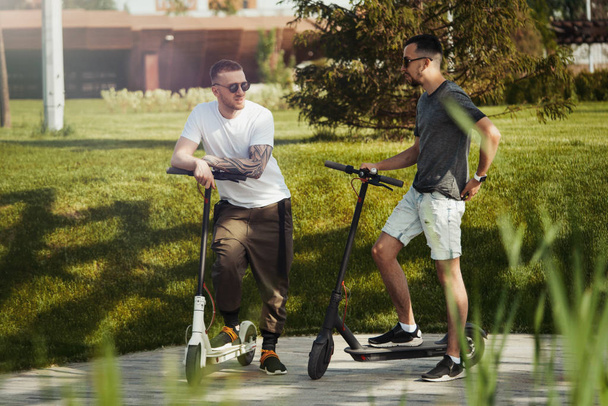 Two handsome men talking next to electric kick scooters at park landscape - Fotó, kép