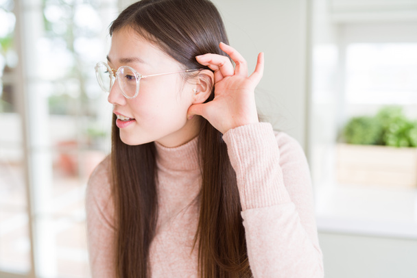 Belle femme asiatique portant des lunettes souriant avec la main sur l'oreille en écoutant une rumeur ou des commérages. Concept de surdité
. - Photo, image