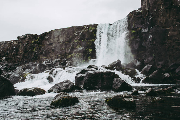 Водопад Оксарарфосс в Национальном парке Тингвеллир, Исландия, текущий из реки в бассейн, наполненный камнями. Холодная облачность весной
 - Фото, изображение