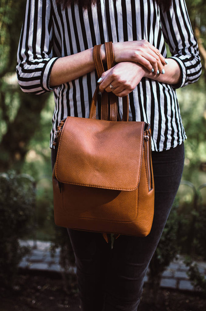 Young female student holding a brown leather backpack in her hands. Stylish girl wearing striped black and white blouse and dark jeans. - Zdjęcie, obraz