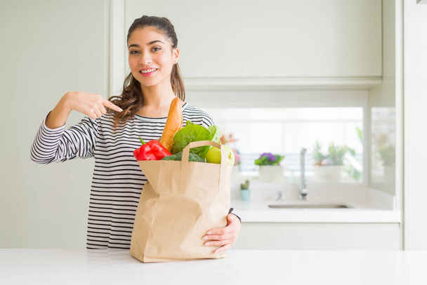 Mladá žena drží papírový sáček plný potravin s překvapenou tváří ukazující prstem na sebe - Fotografie, Obrázek
