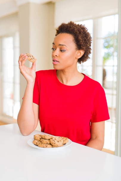 Młoda Afrykańska amerykańska kobieta jedząca ciasteczka czekoladowe z pewnym wyrażeniem na inteligentnym myśleniu twarzy poważnie - Zdjęcie, obraz