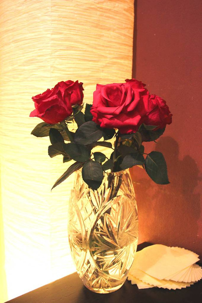 夕方には茶色とベージュの色調でテーブルの上のクリスタルの花瓶の赤いバラの花束 - 写真・画像
