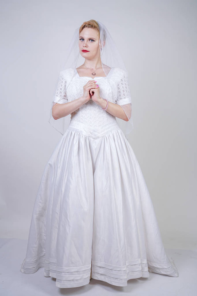 pretty curvy adult woman with short hair wearing long vintage wedding dress with sun style skirt. young caucasian bride with veil on white studio background alone. - Foto, afbeelding