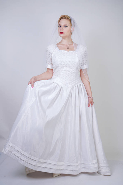 pretty curvy adult woman with short hair wearing long vintage wedding dress with sun style skirt. young caucasian bride with veil on white studio background alone. - Valokuva, kuva