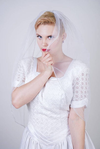 pretty curvy adult woman with short hair wearing long vintage wedding dress with sun style skirt. young caucasian bride with veil on white studio background alone. - 写真・画像
