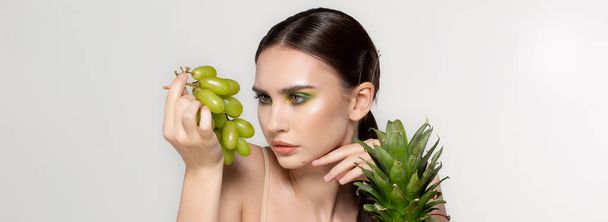 Здорова молода брюнетка дивиться на зелений виноград в руці, фрукти і овочі на столі, студійне фото на сірому фоні
 - Фото, зображення