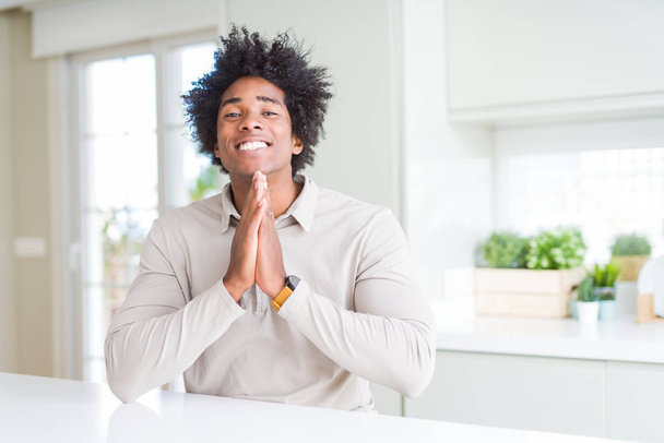 Afrikanisch-amerikanischer Mann betet zu Hause mit den Händen zusammen und bittet um Vergebung, lächelt selbstbewusst. - Foto, Bild