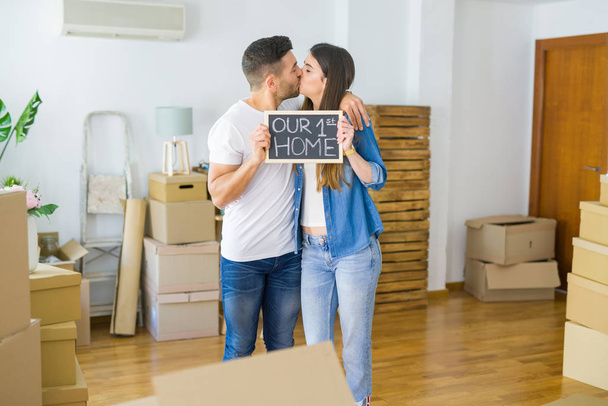 Beautiful young couple moving to a new house, smiling very happy holding blackboard with our first home text - Photo, Image