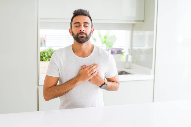 schöner hispanischer Mann lässig weißes T-Shirt zu Hause lächelnd mit den Händen auf der Brust mit geschlossenen Augen und dankbarer Geste im Gesicht. Gesundheitskonzept. - Foto, Bild
