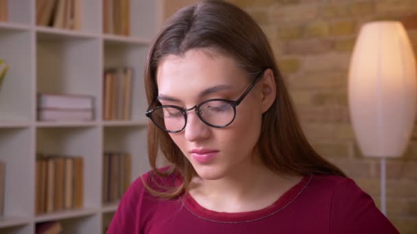 Close-up portrait of young brunette long-haired female student in eye glasses watches calmly into camera. - Кадри, відео