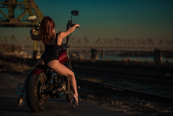 Donna dai capelli rossi in lingerie sexy con i tacchi alti siede su una moto. Attraente ragazza dai capelli rossi siede su una moto al tramonto - Foto, immagini