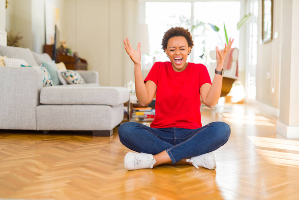Joven mujer afroamericana hermosa sentada en el suelo en casa celebrando loco y loco por el éxito con los brazos levantados y los ojos cerrados gritando emocionado. Concepto ganador
 - Foto, imagen