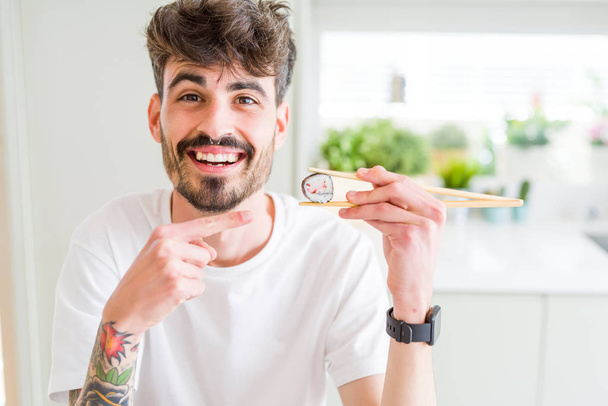 junger Mann isst asiatisches Sushi mit Essstäbchen sehr glücklich zeigt mit Hand und Finger - Foto, Bild