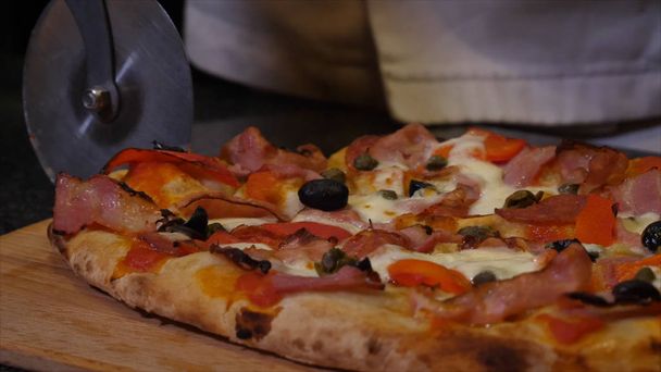 Großaufnahme der Hände des Kochs in schwarzen Schutzhandschuhen, der in der Küche eine frische leckere Pizza mit Speck und Tomaten schneidet. Rahmen. traditionelles italienisches Essen - Foto, Bild