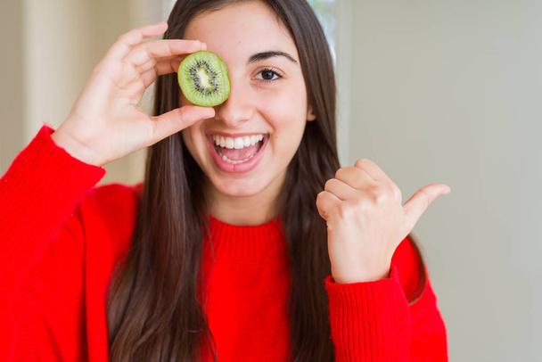 Kaunis nuori nainen syö puoli tuoretta vihreää kiiviä osoittaen ja osoittaen peukalolla sivulle hymyillen
 - Valokuva, kuva