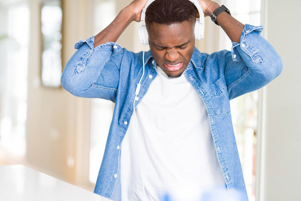 Афроамериканец в наушниках слушает музыку, страдающую от головной боли в отчаянии и стрессе из-за боли и мигрени. Руки на голову
. - Фото, изображение