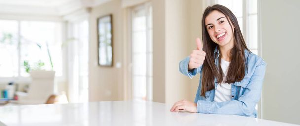 Широке кутове зображення красивої молодої жінки, яка сидить на білому столі вдома, роблячи щасливі великі пальці вгору жестом рукою. Затвердження виразу, який показує успіх фотоапарата
. - Фото, зображення