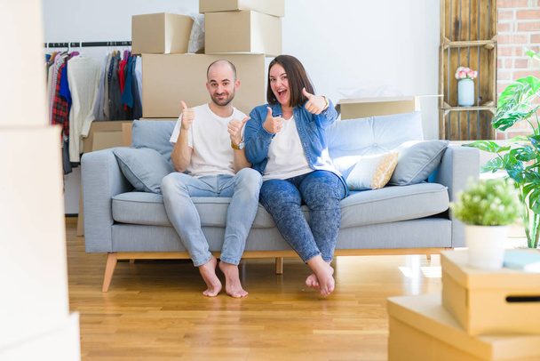 Mladý pár sedící na pohovce arround lepenkové krabice pohybující se k novému domu úspěch znamení dělá pozitivní gesto rukou, palce nahoru s úsměvem a šťastný. Veselý výraz a gesto vítěze. - Fotografie, Obrázek