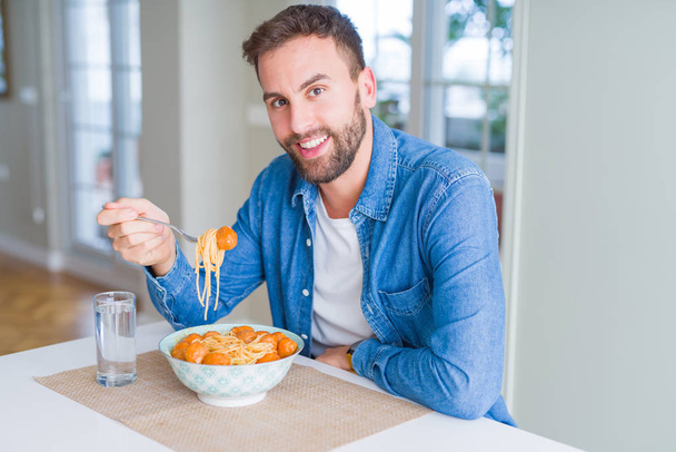 Hombre guapo comiendo pasta con albóndigas y salsa de tomate en casa mientras sonríe a la cámara
 - Foto, imagen
