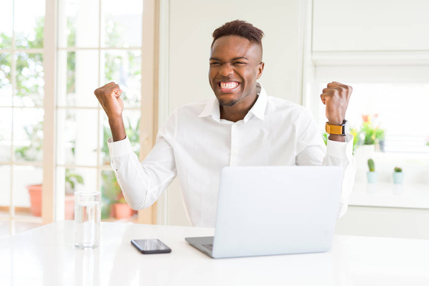Hombre de negocios afroamericano que trabaja con computadora portátil celebrando sorprendido y sorprendido por el éxito con los brazos levantados y los ojos abiertos. Concepto ganador
. - Foto, Imagen