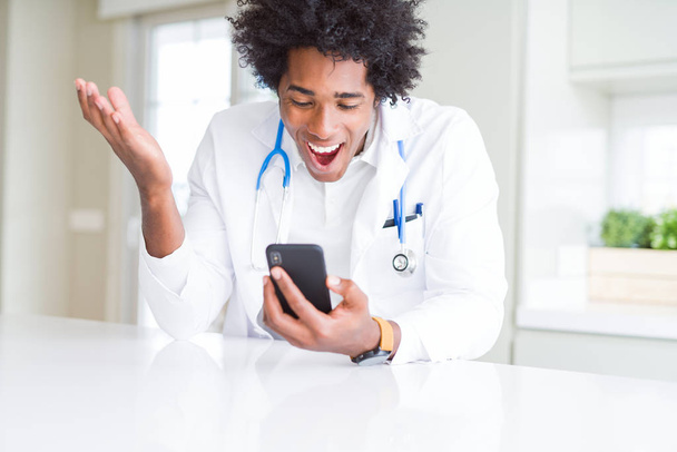African American doctor man using smartphone at the clinic very happy and excited, winner expression celebrating victory screaming with big smile and raised hands - Фото, зображення